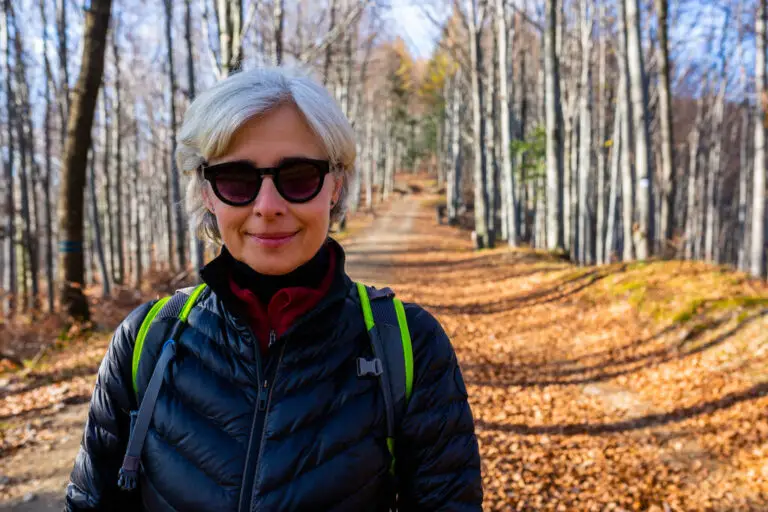 senior woman wearing sunglasses and a backpack enjoys a peaceful hike on a forest trail during autumn.