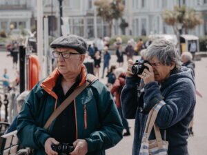 Senior couple with cameras taking photos on a sunny seaside promenade lined with palm trees and white buildings.