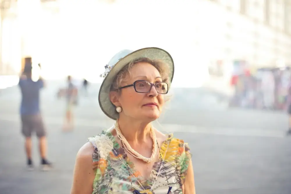 Elderly woman in a hat and floral dress gazes thoughtfully, set against a blurred street background. A serene moment in one of the best vacation destinations for single seniors