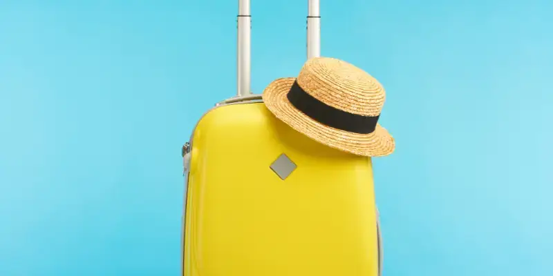 Bright yellow suitcase with an extended handle and a straw hat on top against a blue background, symbolizing adventure. Best luggage for solo travelers seeking convenience.
