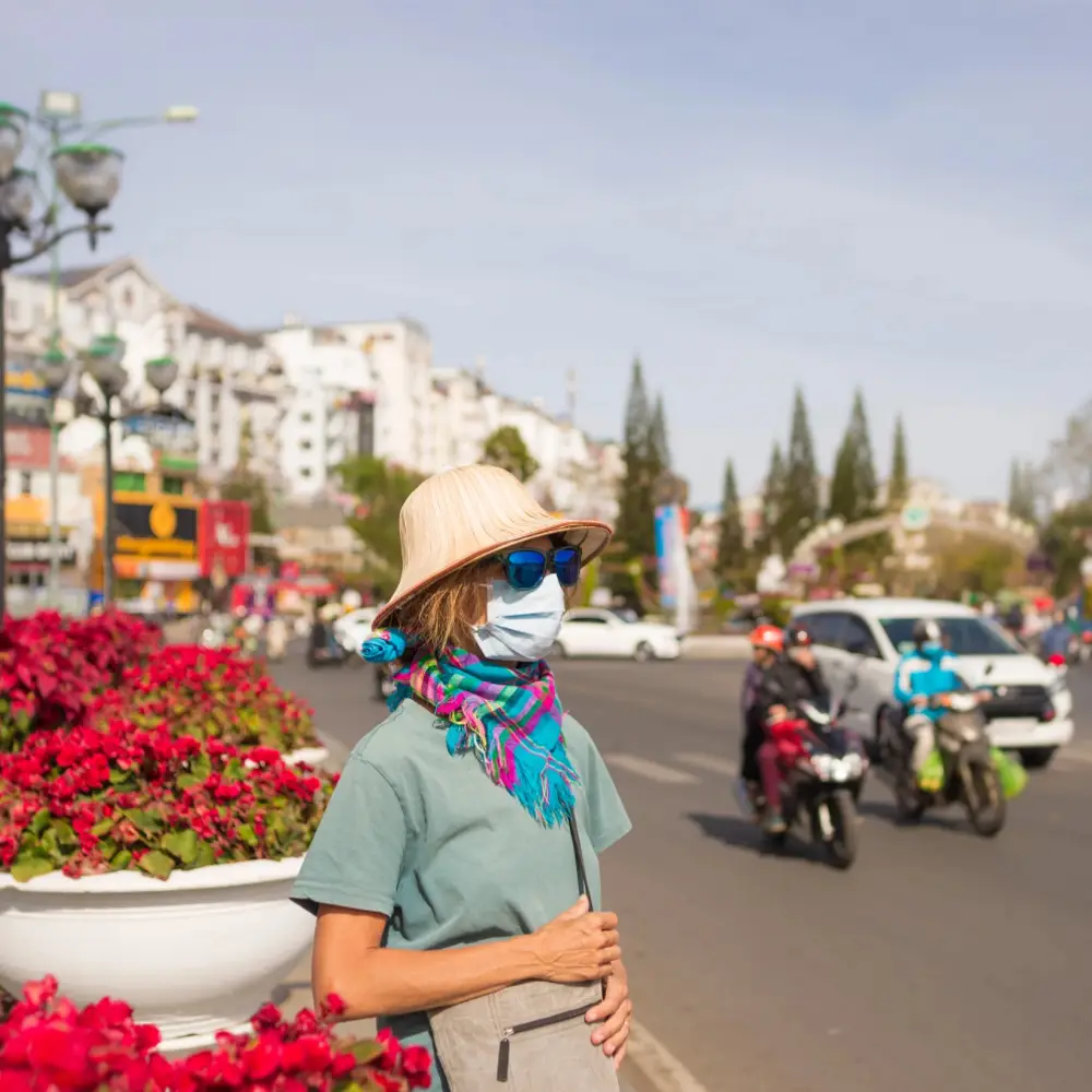 A tourist in Vietnam wearing a hat, sunglasses, and a mask stands beside a busy city street with red flowers, holding a bag as cars and motorcycles pass by.
