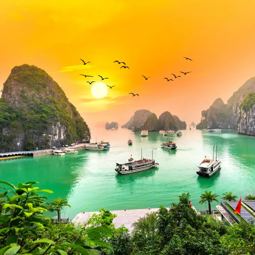 Best time to visit Vietnam, Sunset over a scenic bay with limestone karsts, emerald-green water, and boats, as birds soar under an orange sky.