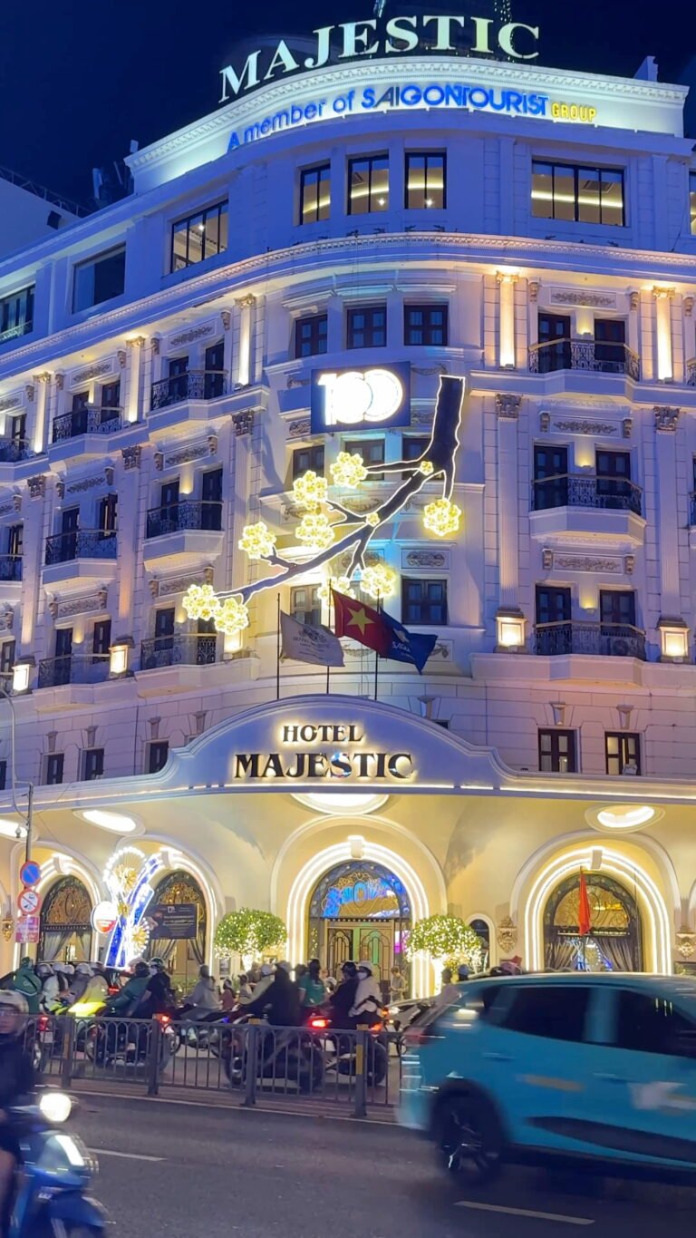 The historic Hotel Majestic in Ho Chi Minh City, Vietnam, illuminated at night with ornate French colonial architecture, festive light decorations, and motorbikes streaming past on the street below.