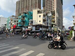 A busy street intersection in Ho Chi Minh City, Vietnam, showing dozens of motorbikes and pedestrians crossing mid-traffic, with colorful shophouses and modern high-rise construction in the background.