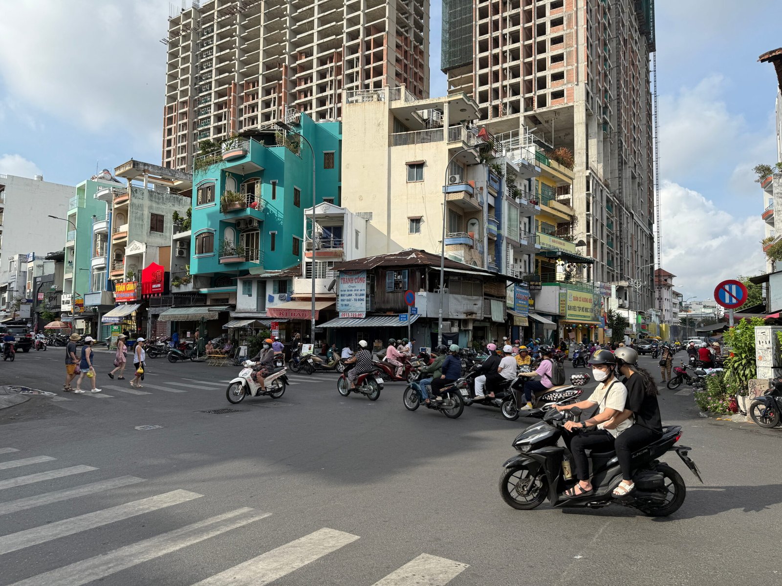 A busy street intersection in Ho Chi Minh City, Vietnam, showing dozens of motorbikes and pedestrians crossing mid-traffic, with colorful shophouses and modern high-rise construction in the background.