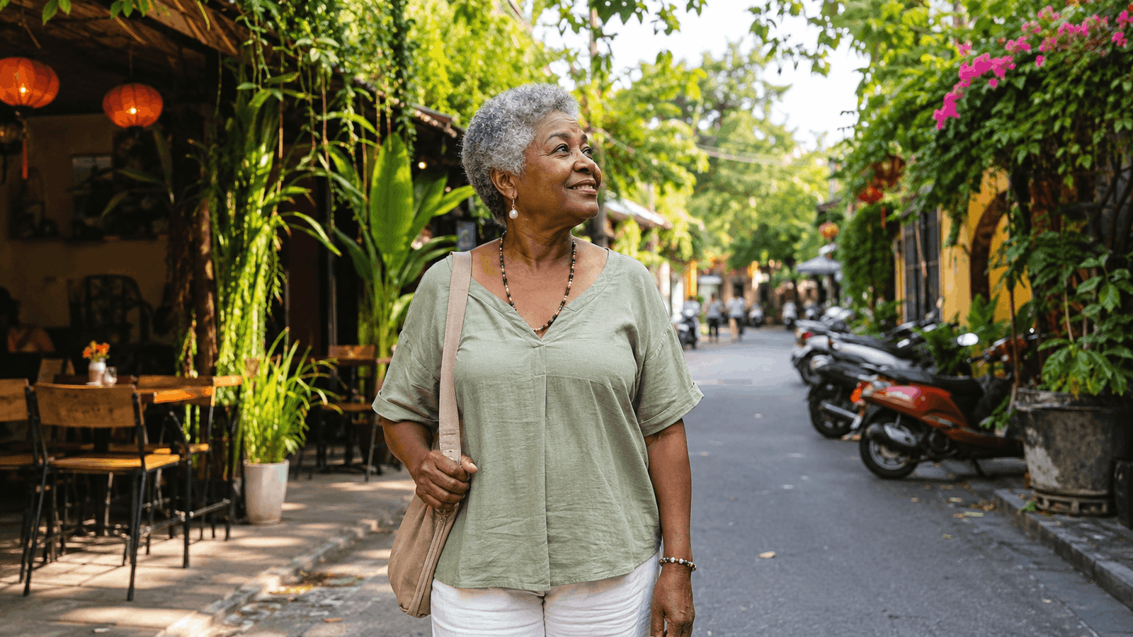 African American senior woman walking along a leafy street in Southeast Asia, with cafes, tropical plants, and motorbikes in the background.